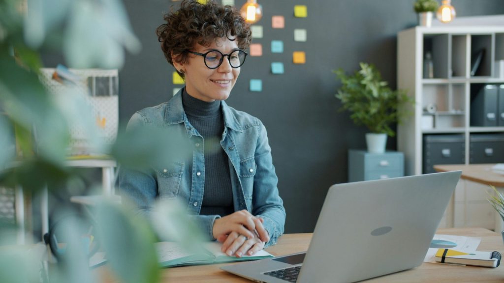 A smiling woman with curly hair works at a laptop
